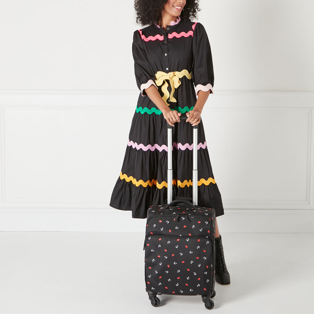 A woman in a black and colourful dress stands holding a small black rolling suitcase with a repeating pattern of red lips. The background is white.
