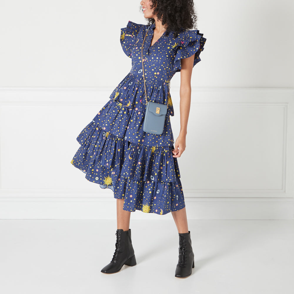 Woman in a blue celestial print dress with ruffled skirt and a blue crossbody bag. She's wearing black lace-up boots. The background is white.
