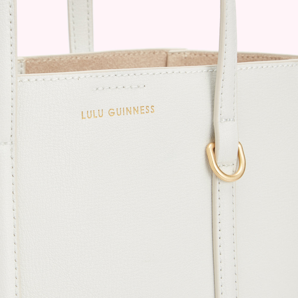 Close-up of a white Lulu Guinness tote bag showing the brand's logo and gold-toned hardware against a light pink background.
