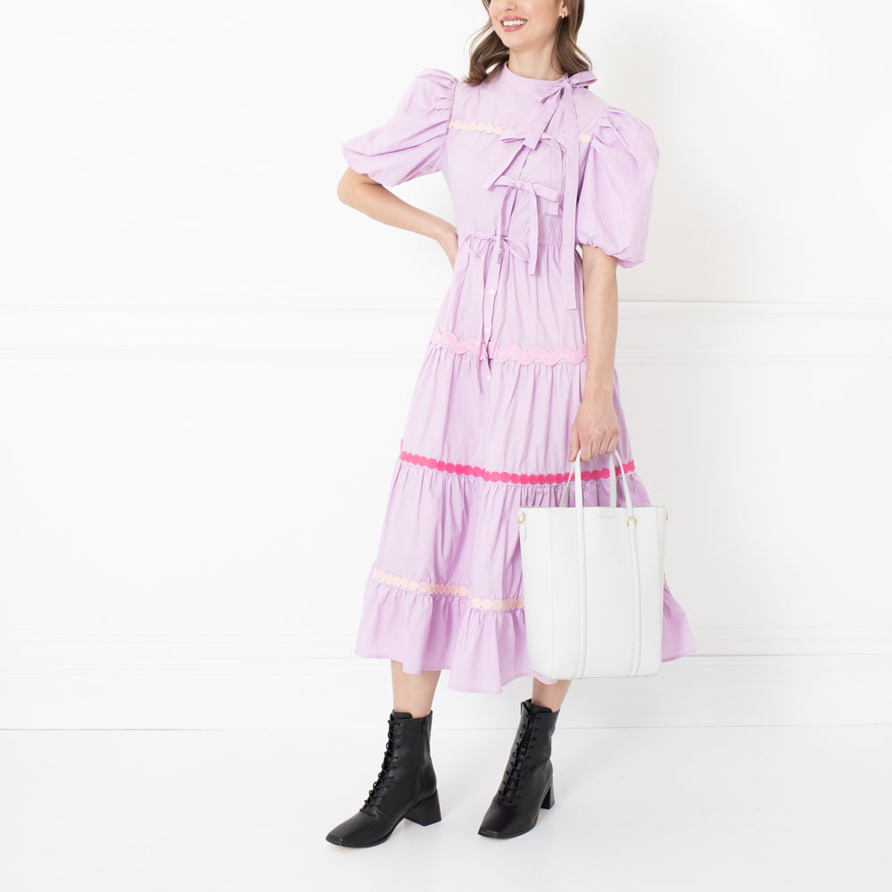 Woman in lavender tiered midi dress with puff sleeves, white tote bag, and black ankle boots against a white background.
