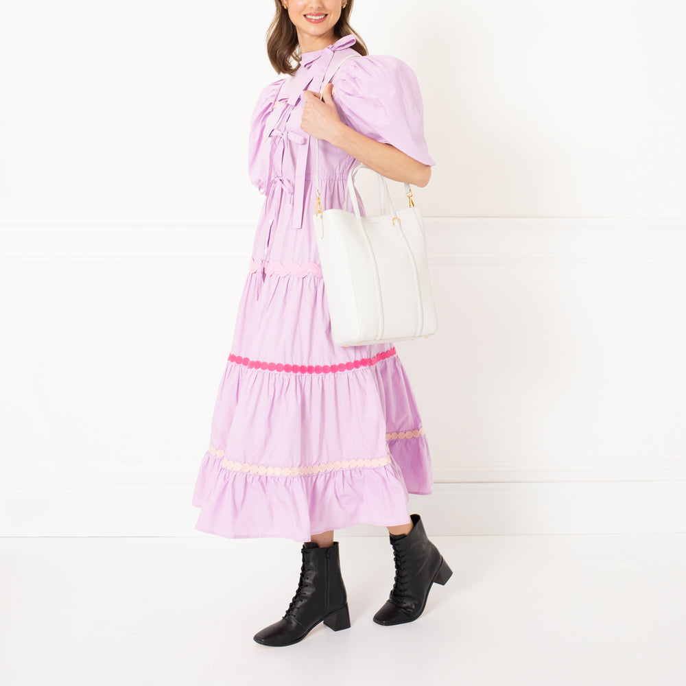 Woman in lavender dress carrying a white tote bag and wearing black ankle boots against a white background.
