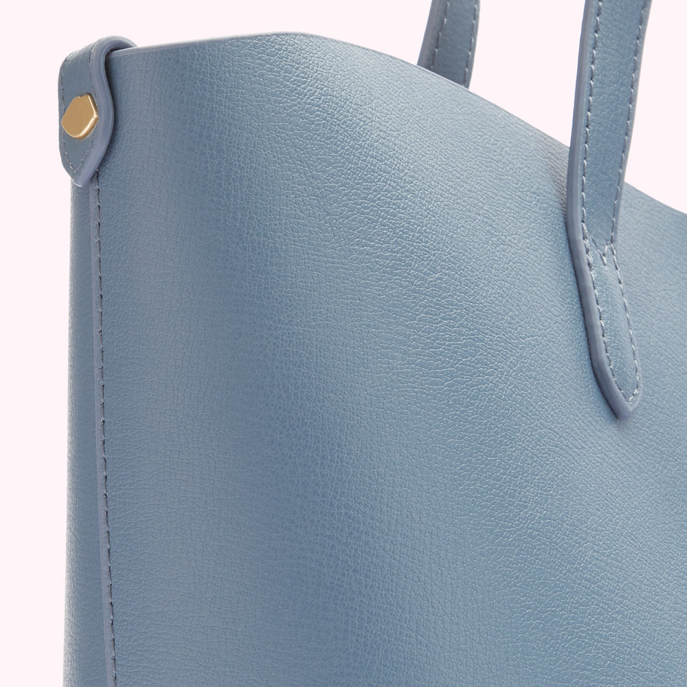 Close-up of a dusty blue tote bag's textured leather, showcasing its stitching and gold-toned hardware.
