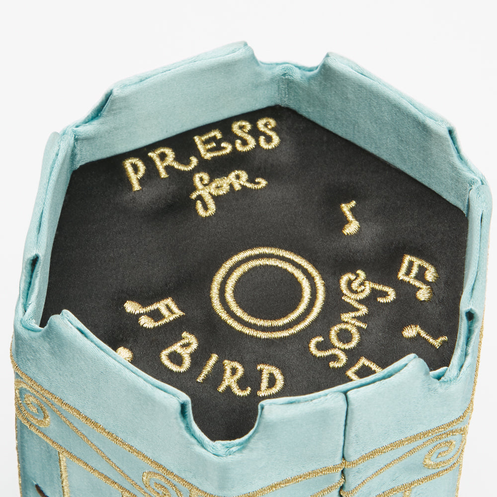 Close-up of the base of a light blue birdcage shaped handbag with gold embroidery that reads "Press for Bird Song" and musical notes.