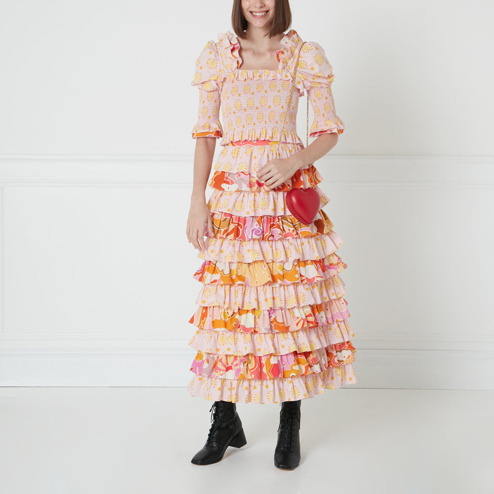 A woman in a pink and orange tiered maxi dress and black ankle boots holds a small red heart-shaped purse.
