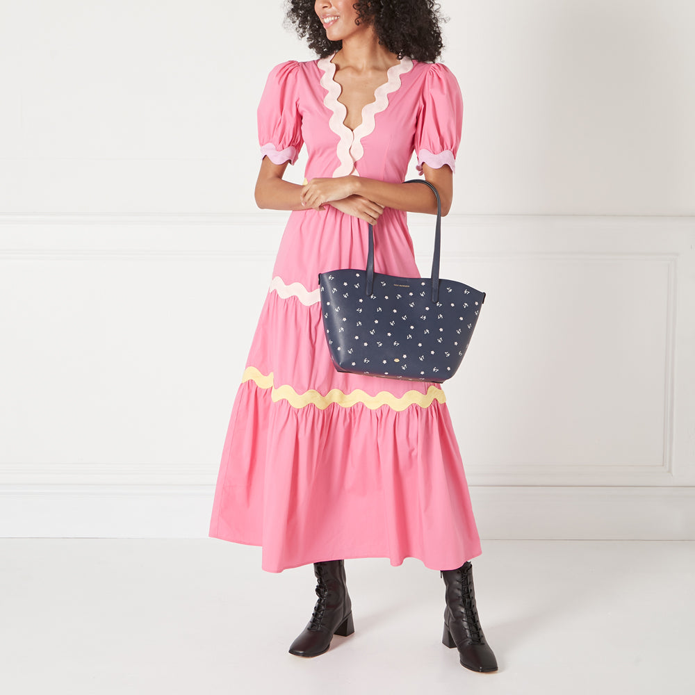 A woman in a pink dress holds a navy blue tote bag with a small white floral print against a white background.

