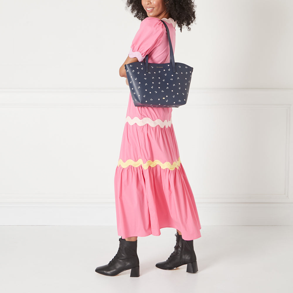 A woman in a pink dress carries a navy tote bag with a floral pattern against a white background.