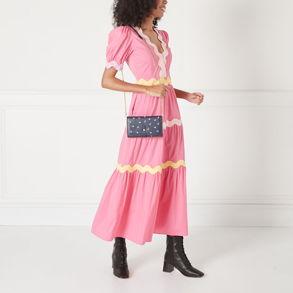A woman in a pink dress carries a navy blue crossbody wallet with a gold chain strap. The wallet has small white floral details.
