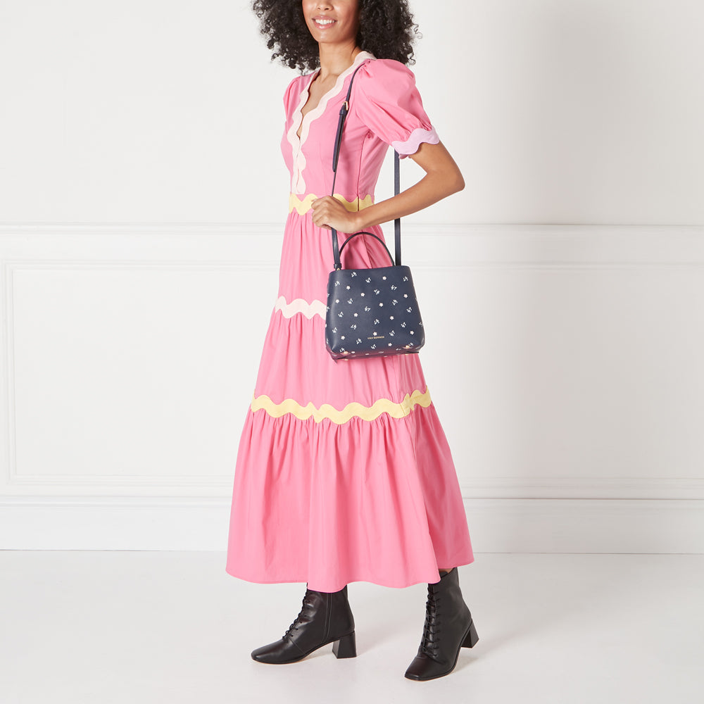 A woman in a pink dress carries a navy blue crossbody bag with a floral pattern. The background is white.
