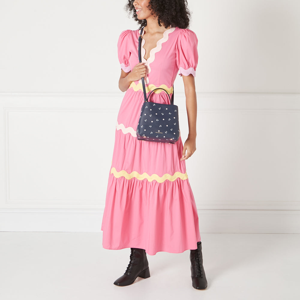Woman in pink dress holding a navy blue crossbody bag with a floral pattern against a white background.