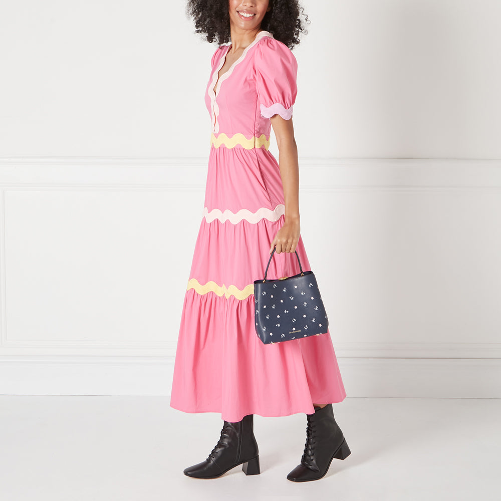 A woman in a pink dress holds a navy blue handbag with a white floral pattern against a white background.
