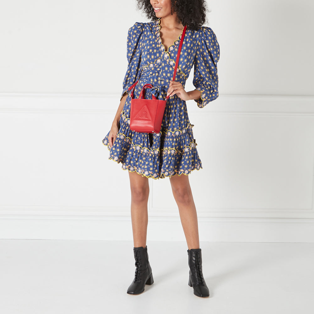 A woman in a blue floral dress and black boots carries a small red crossbody bag.
