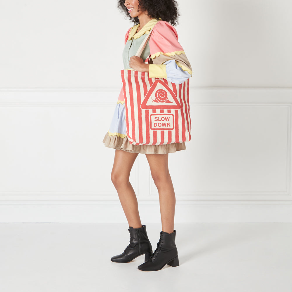 A woman in a pastel dress carries a red and white striped tote bag that says "Slow Down" in a vintage-style traffic sign.