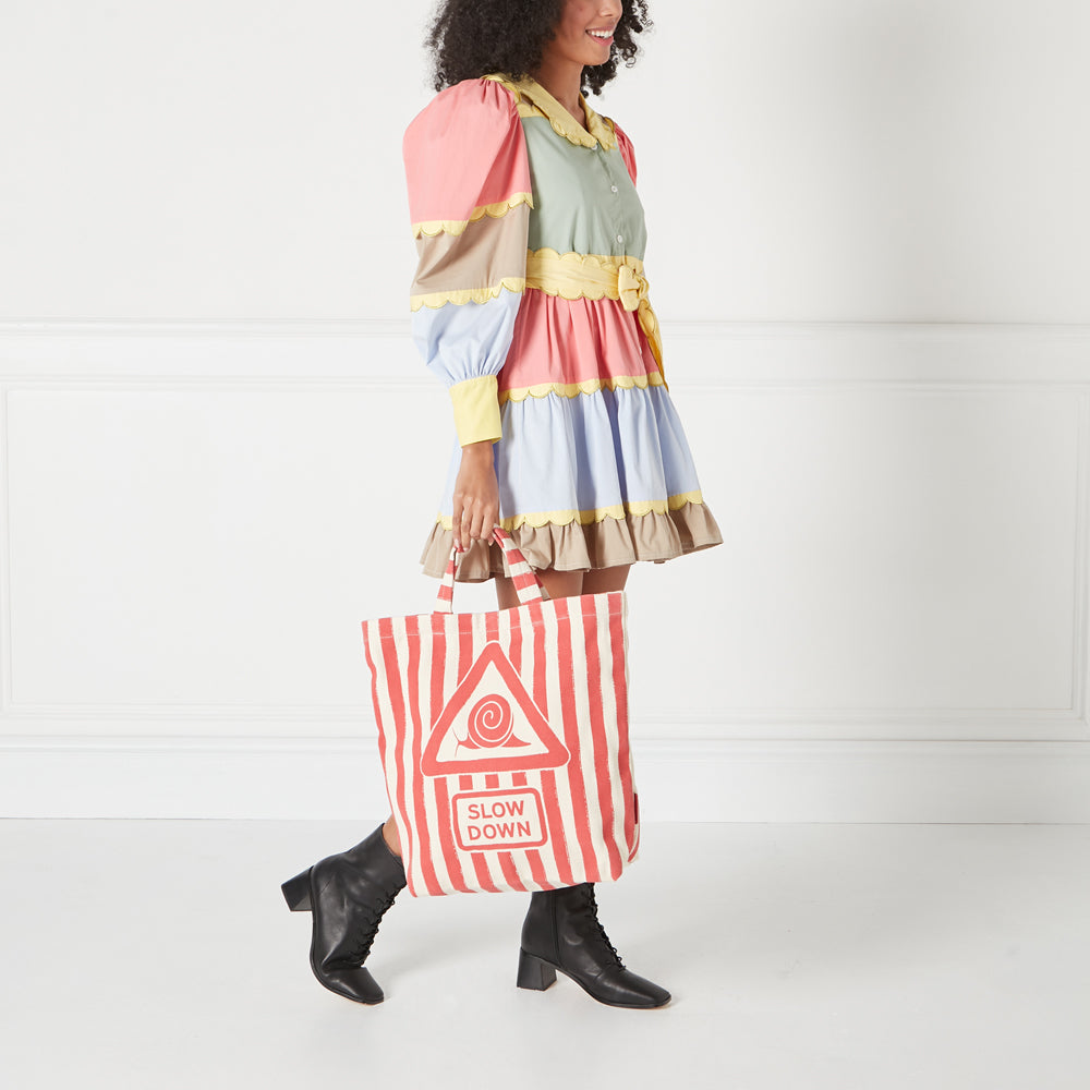 Woman in a pastel coloured dress carrying a red and white striped tote bag with a snail and "Slow Down" printed on it.
