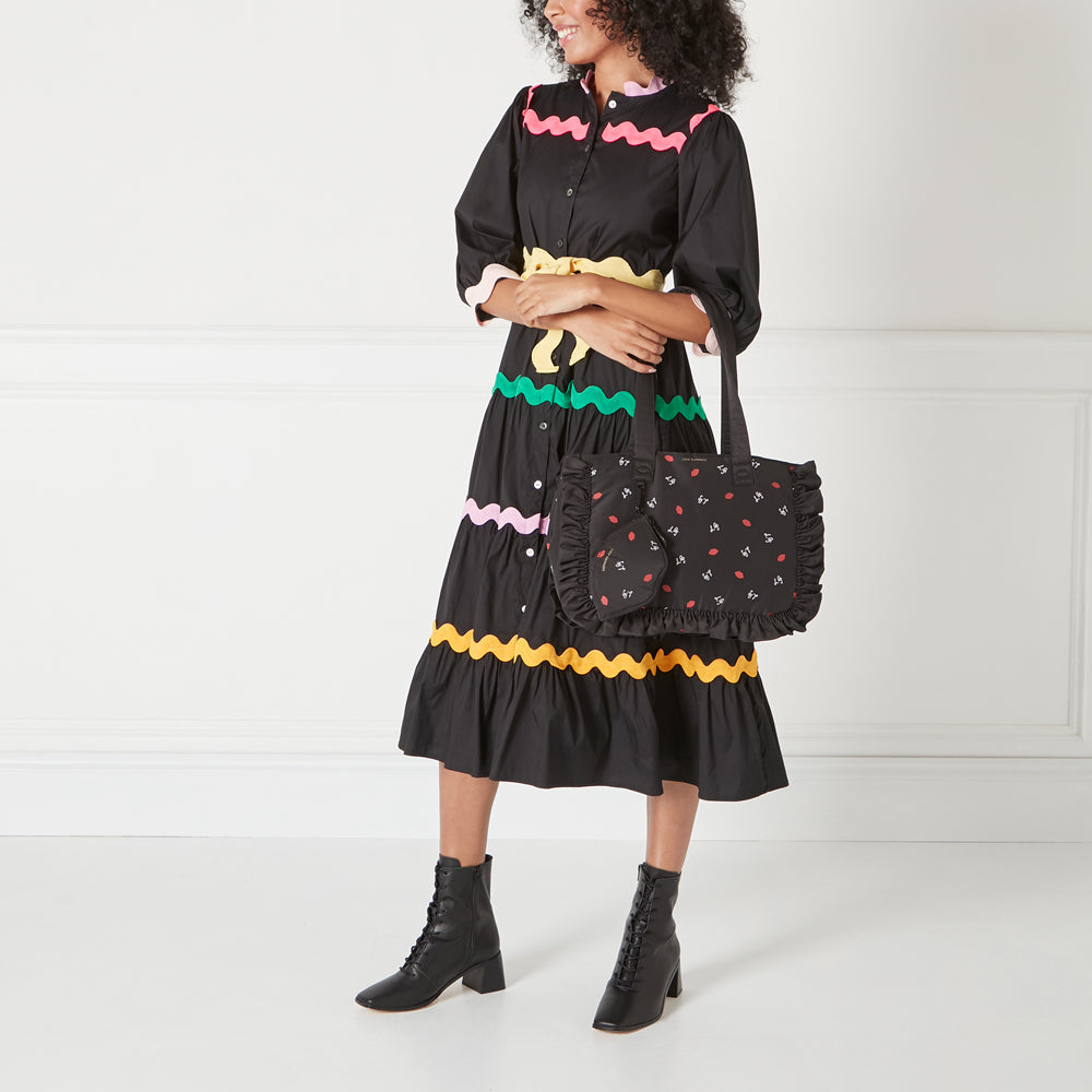 A model wears a black dress with colourful scalloped trim and black lace-up boots. She carries a black tote bag with a ruffled trim and a small matching pouch. The bag is decorated with a pattern of lips.
