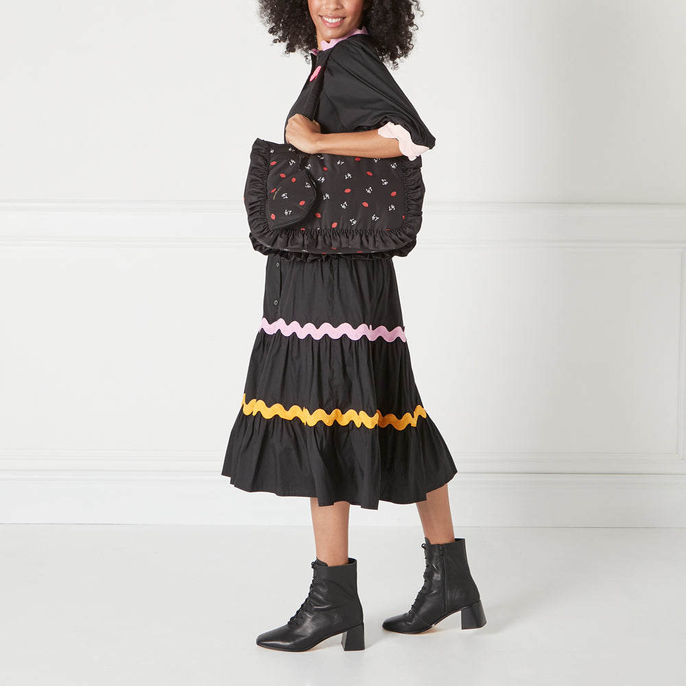 Woman in black dress and boots holding a black ruffled tote bag with a red lips print against a white background.