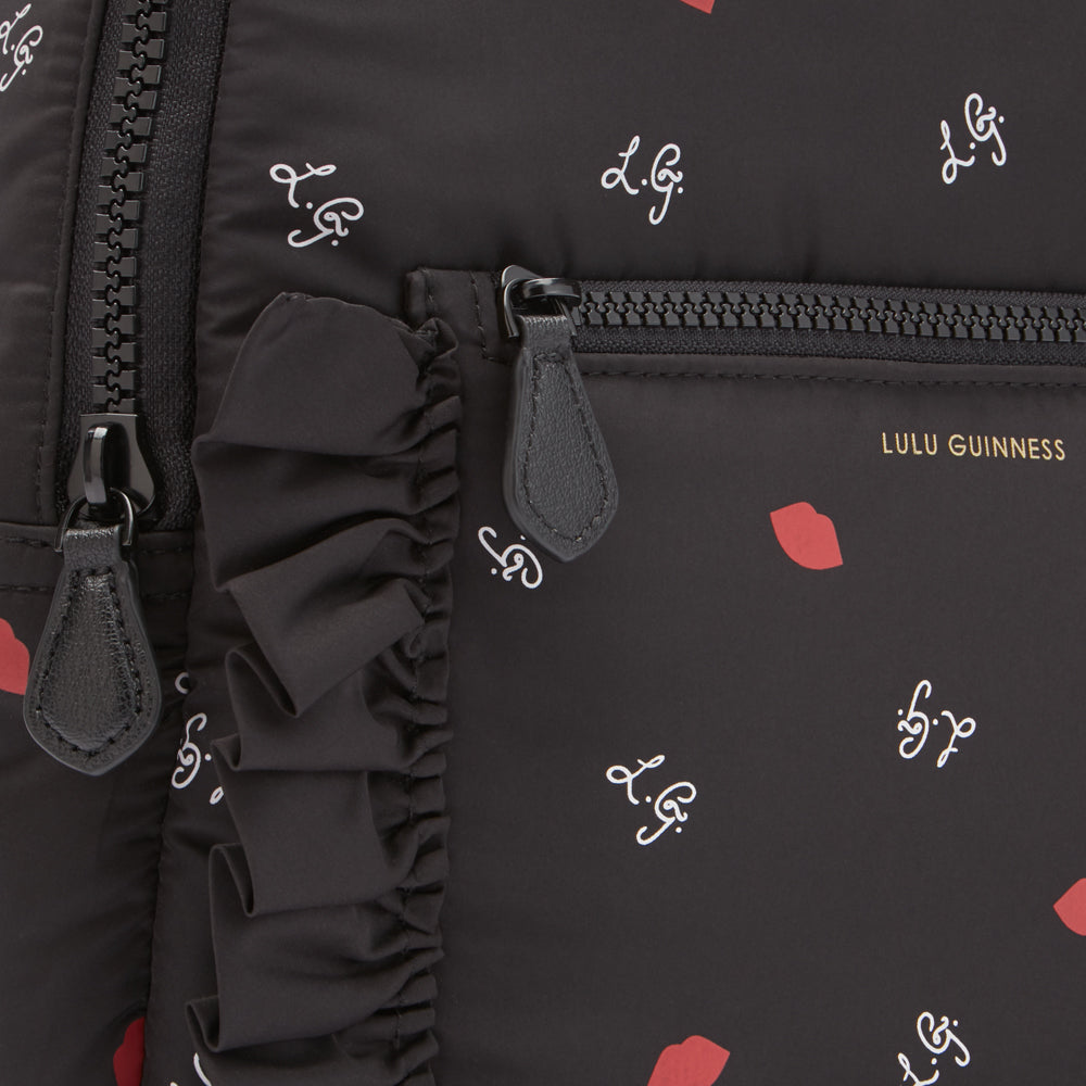 Close-up of a black Lulu Guinness backpack with a ruffled detail, featuring a repeating pattern of red lips and the brand's logo.
