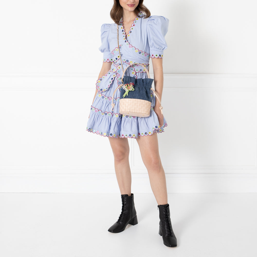 A woman in a light blue dress and black ankle boots carries a small woven bag with a denim top. The background is white.
