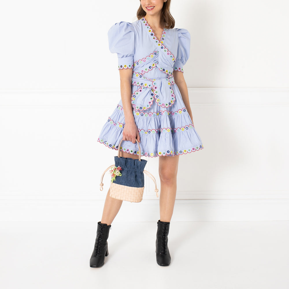 A woman in a light blue embroidered dress and black ankle boots holds a small woven handbag with floral accents against a white background.

