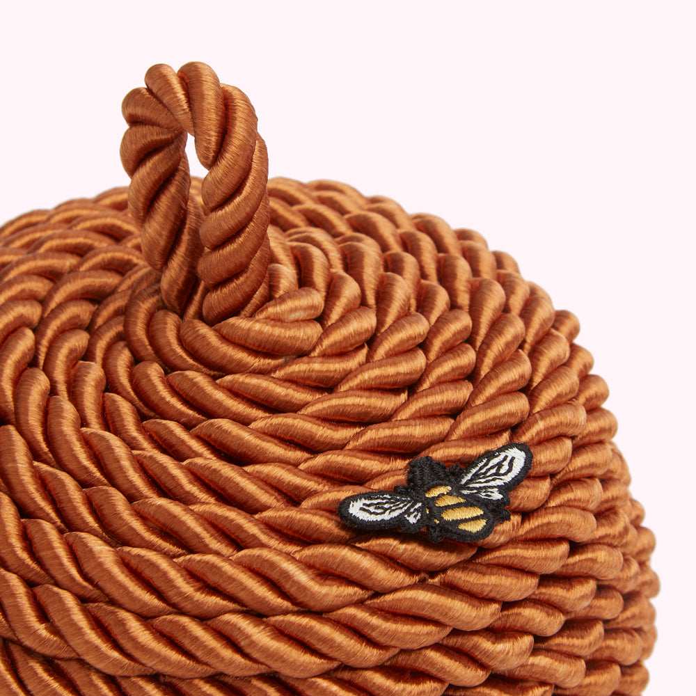 Close-up of a burnt orange, rope-constructed beehive-shaped object with a small embroidered bee on its side.
