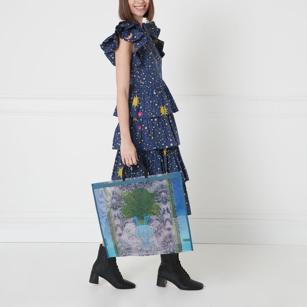 Woman in a navy blue tiered dress with a celestial print holding a large mesh tote bag with a floral print against a white background.
