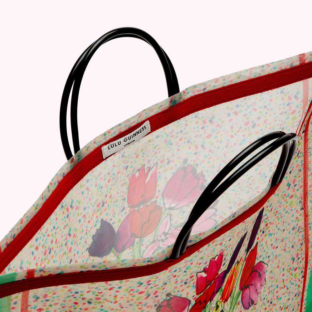 Close-up view of the inside of a Lulu Guinness London tote bag. The bag is a sheer fabric with a multicolored confetti print and a tulip design. The handles are black. The lining is red.
