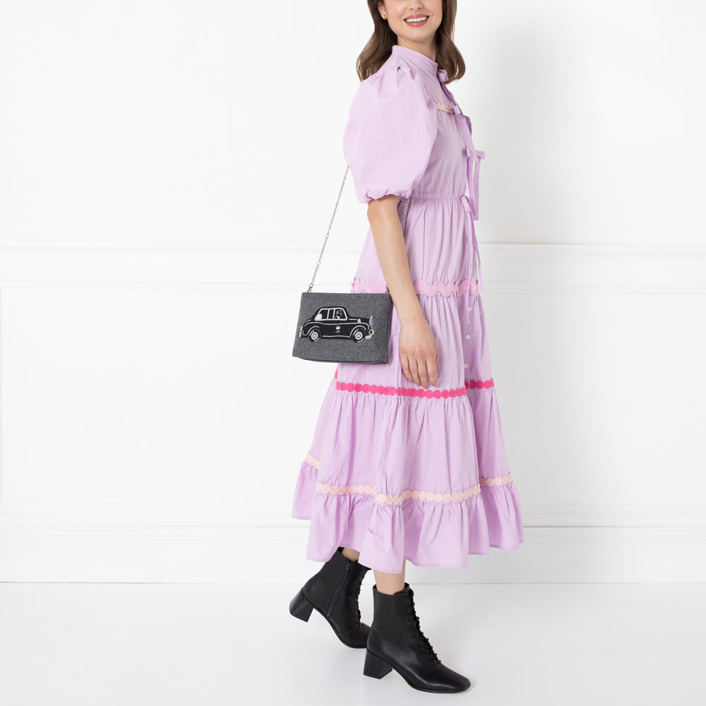 Woman in lavender dress and black ankle boots carrying a grey crossbody bag with a black cab embroidered on it.