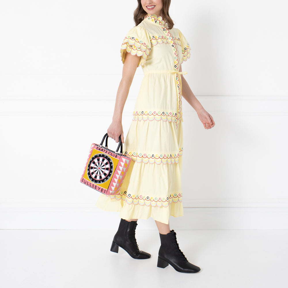 A woman in a yellow embroidered dress carries a pink and yellow darts-themed handbag against a white background.
