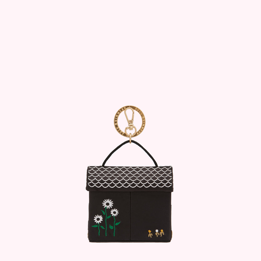 Black and white house-shaped keychain with gold ring, featuring embroidered flowers and small figures.
