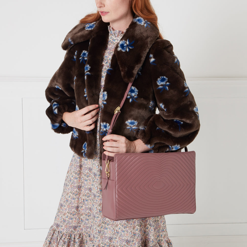 Woman in brown floral faux fur jacket and floral dress holding a dusty rose quilted crossbody bag against a white background.
