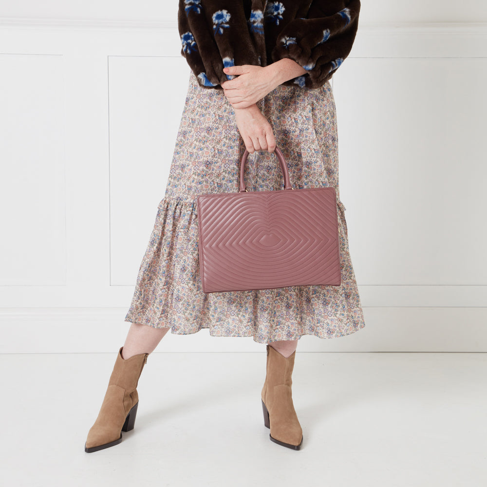 Woman holding a mauve quilted tote bag, wearing a floral midi skirt and brown boots.
