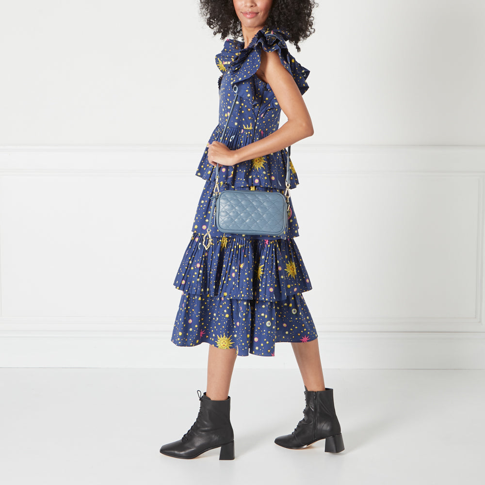 A model wearing a blue dress and black ankle boots carries a quilted blue crossbody bag against a white background.
