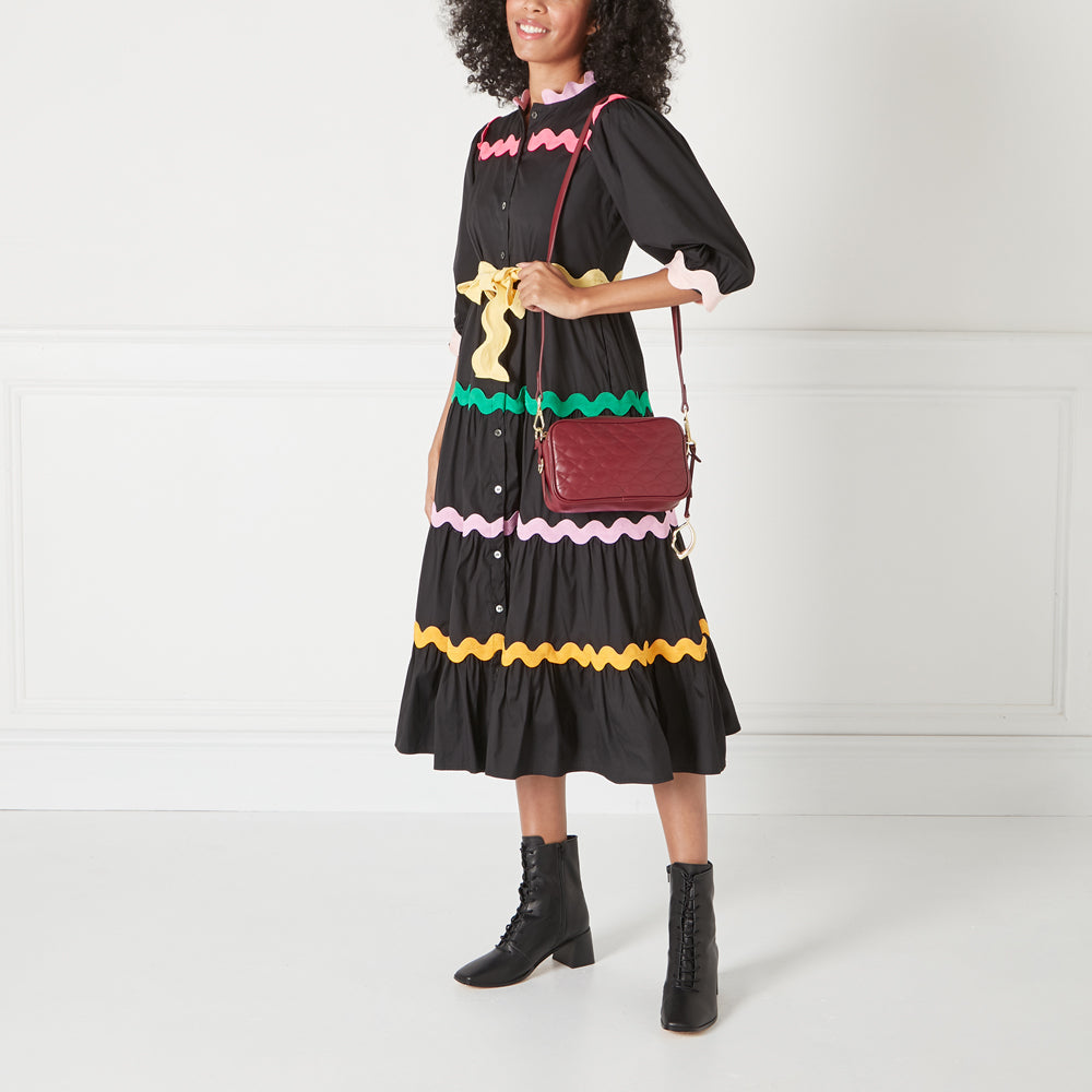 Woman in a black dress with colourful trim carries a burgundy crossbody bag.
