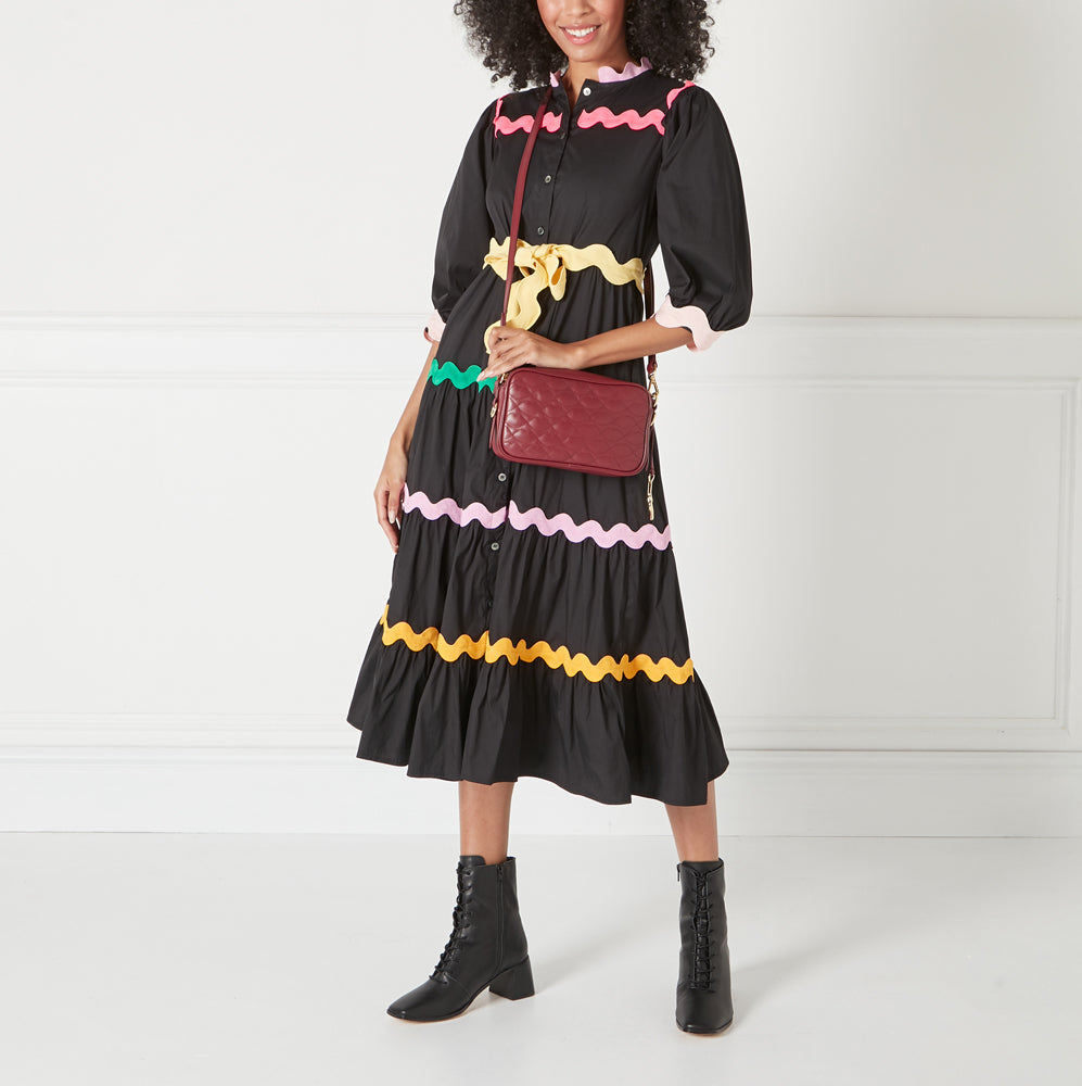 A woman in a black dress with colourful trim carries a burgundy quilted crossbody bag.
