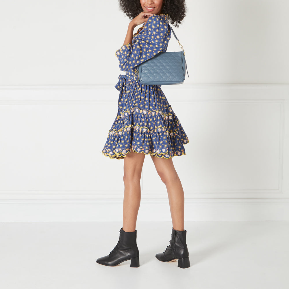 A woman in a blue floral dress and black ankle boots carries a blue quilted shoulder bag against a white background.
