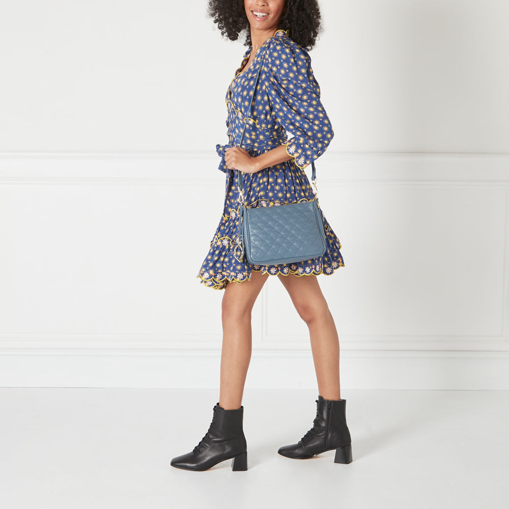 Woman in a blue floral dress and black ankle boots carrying a quilted blue crossbody bag against a white background.