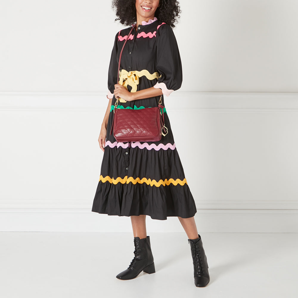 A woman in a black dress with colourful trim carries a quilted burgundy crossbody bag and wears black boots.