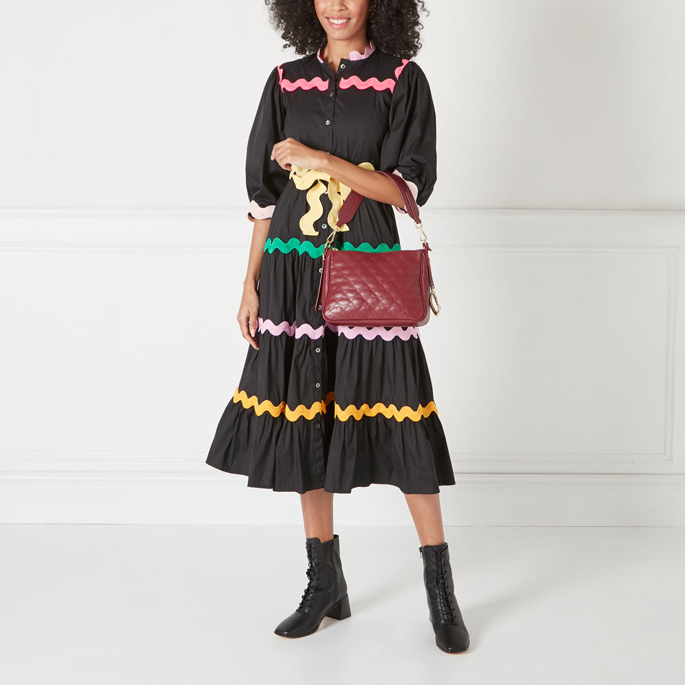 A woman in a black dress with colourful trim carries a burgundy quilted crossbody bag.
