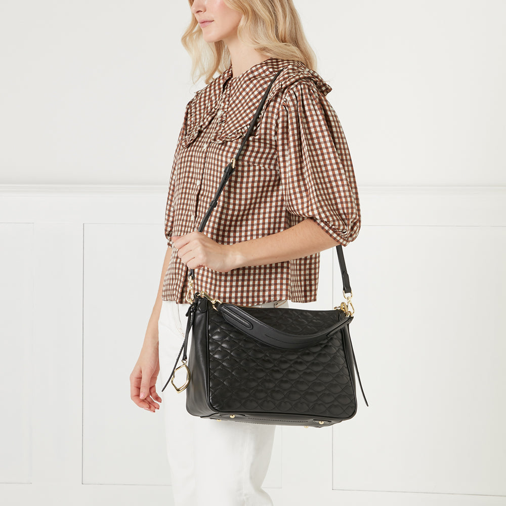 Woman in brown gingham shirt holding black quilted crossbody bag against white background.
