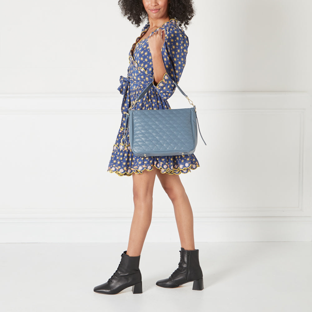 A woman in a blue floral dress carries a quilted blue shoulder bag and wears black ankle boots against a white background.
