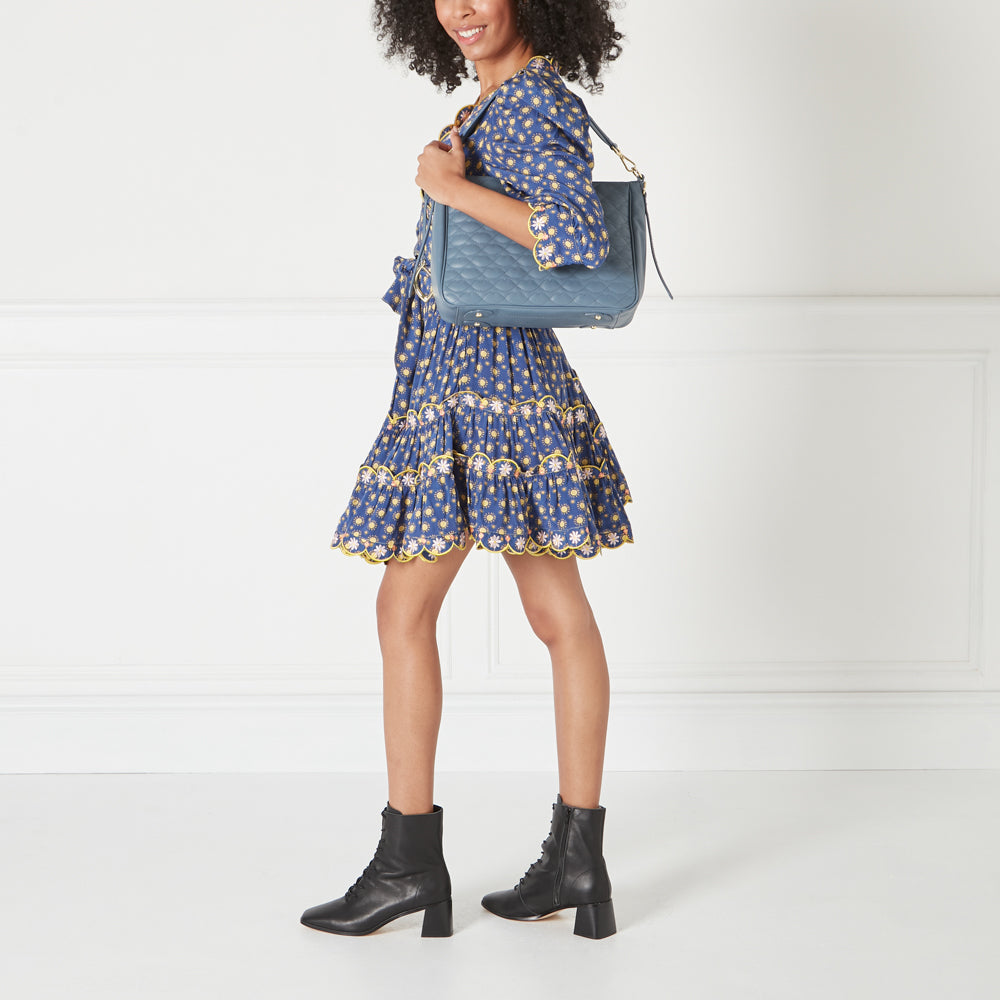 A woman in a floral dress and black ankle boots carries a quilted blue shoulder bag against a white background.
