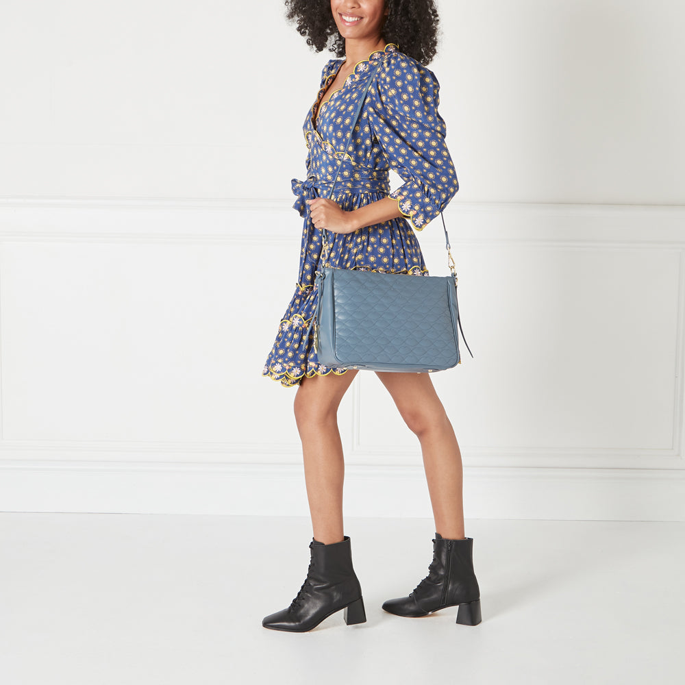 Woman in blue floral dress and black ankle boots carries a blue quilted shoulder bag against a white background.
