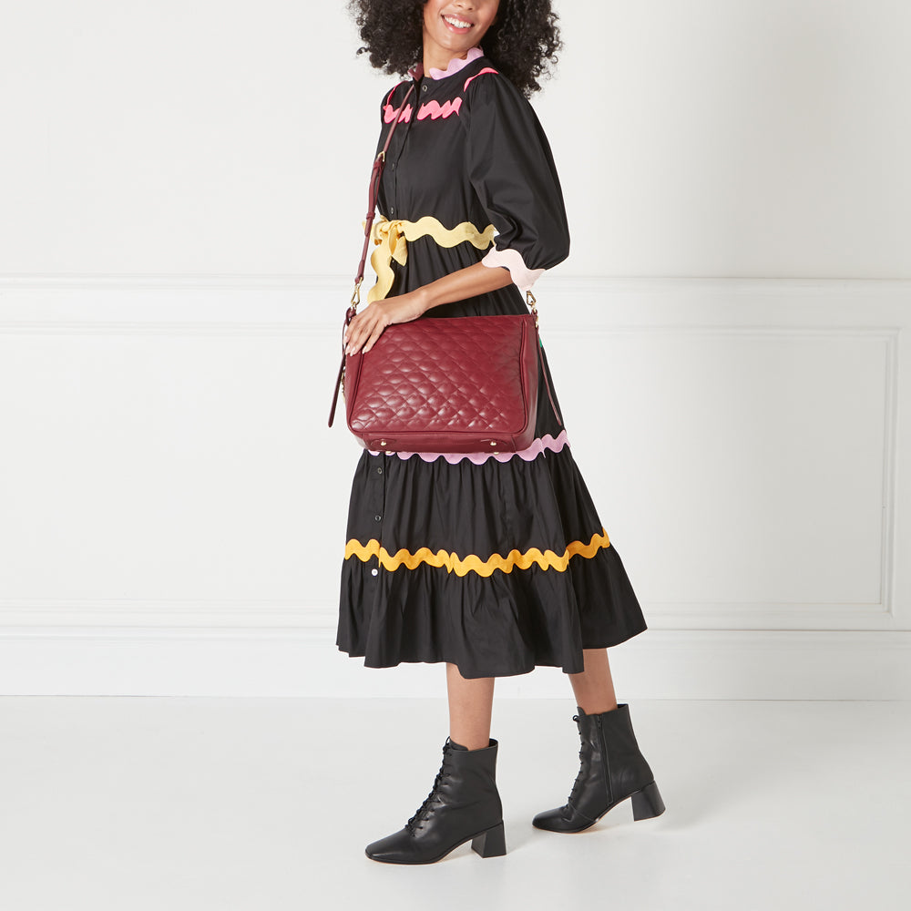 A woman in a black dress with colourful trim carries a burgundy quilted handbag.
