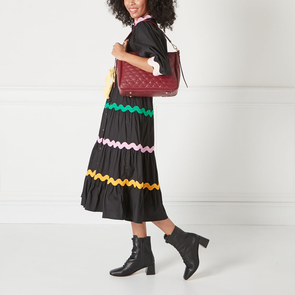 A woman in a black dress with colourful trim carries a burgundy quilted handbag.
