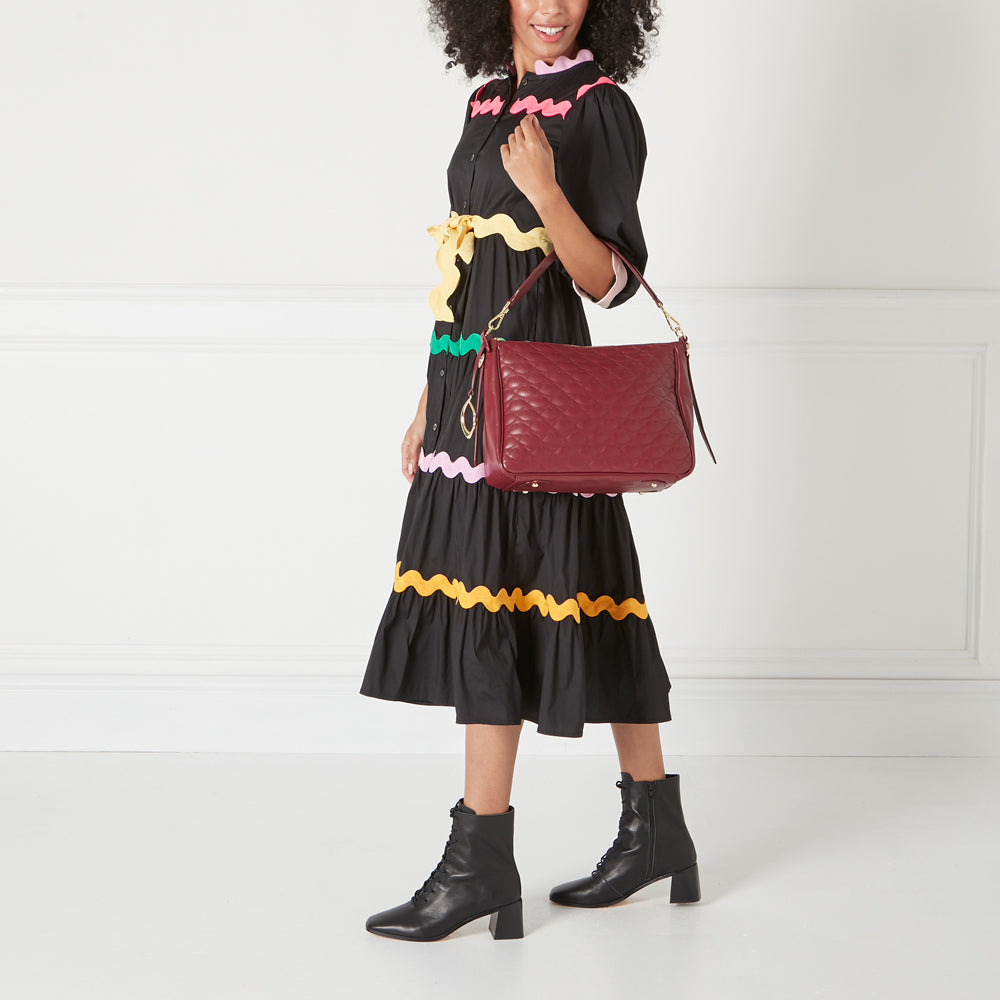 Woman in black dress holding a maroon quilted shoulder bag against a white background.
