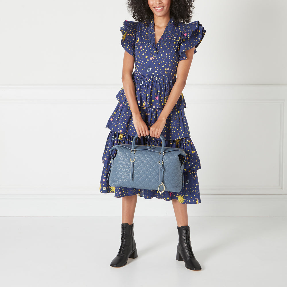 A woman in a navy blue dress with celestial print holds a quilted blue satchel bag.

