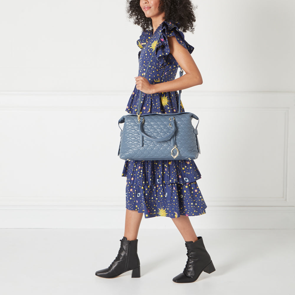 A woman in a blue dress and black boots carries a quilted blue leather handbag. The background is white.
