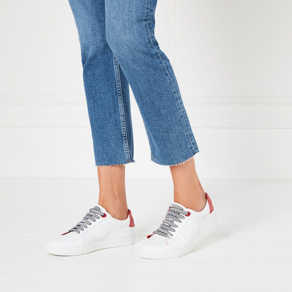 White leather trainers with navy and white striped laces and a red heel detail, modeled on a person wearing cropped blue jeans against a white background.
