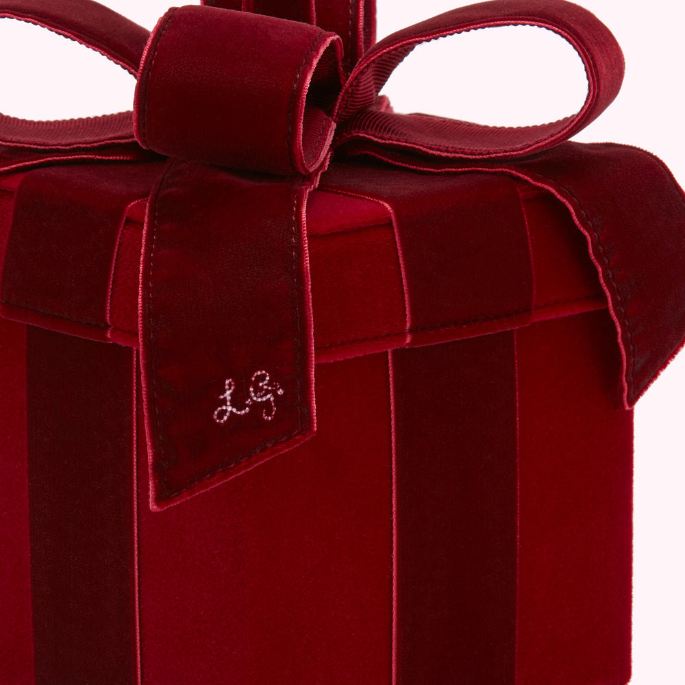 Close-up of a dark red velvet gift box with a large bow; the box has vertical stripes and the initials "LG" are embroidered on the front.
