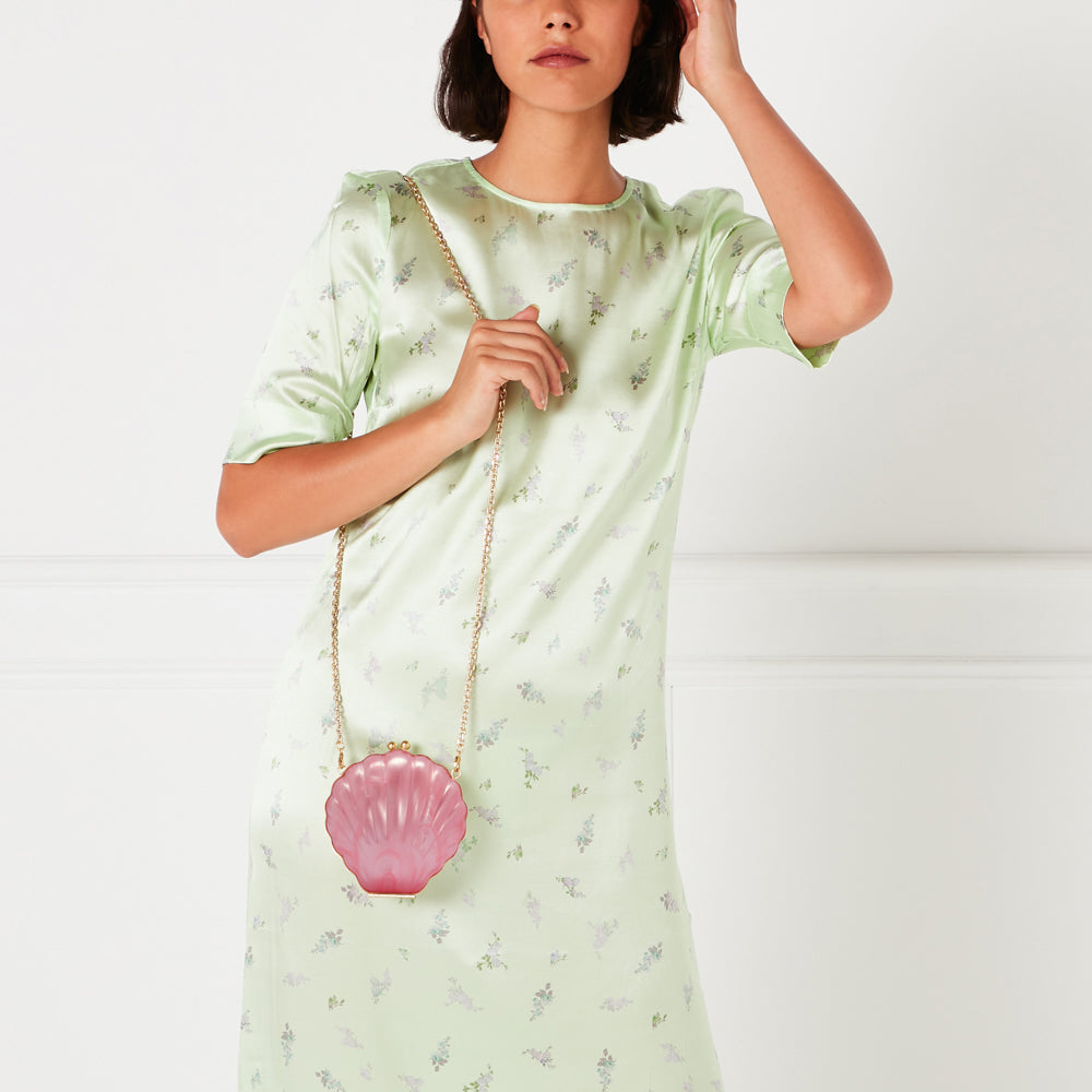Woman in a light green floral dress holding a pink seashell-shaped purse with a gold chain strap.
