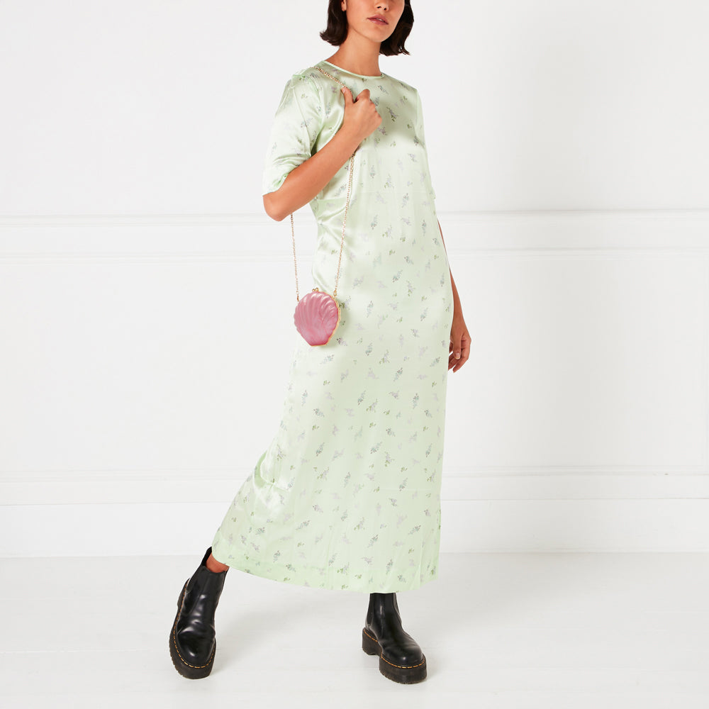Woman in a light green floral maxi dress and black Chelsea boots, carrying a pink shell-shaped purse. The background is white.
